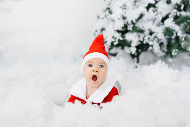 Vauva pienessä joulupukin asussa ja hatussa makaa selällään keinotekoisessa lumessa. Poika on yllättynyt myynnistä. Joululomien käsite. Tervehdyskortti
 - Valokuva, kuva