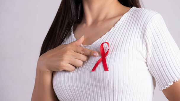 Closeup of woman hand pointing to red badge ribbon on chest to support AIDS Day. Healthcare, medicine and AIDS awareness concept. - 写真・画像