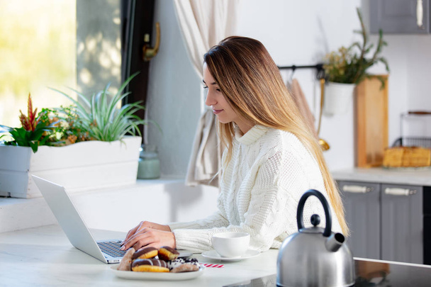 Nuori nainen työskentelee tietokoneella hir keittiö ja juo teetä
 - Valokuva, kuva