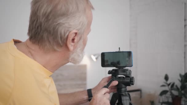 παππούς και την τεχνολογία, ηλικιωμένοι άνδρες blogger είναι επιρροή έτοιμος να χρησιμοποιήσει το έξυπνο τηλέφωνό του για την καταγραφή vlog ρυθμίσεις κάμερα στη συνέχεια εισέρχεται στο πλαίσιο στο δωμάτιό του για τη λήψη περιεχομένου - Πλάνα, βίντεο