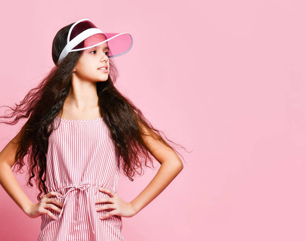 Menina adolescente bonita em um boné e vestido listrado rosa alegremente mostra um sinal vencedor rindo sorte em um fundo rosa
  - Foto, Imagem