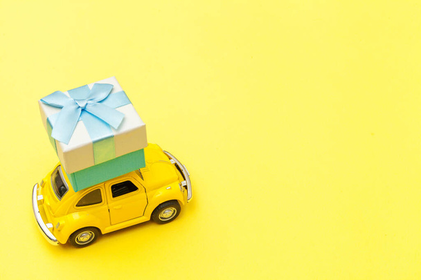 Simply design yellow vintage retro toy car delivering gift box on roof isolated on trendy yellow background. Christmas New Year birthday Valentine's day celebration present romantic concept - Fotó, kép