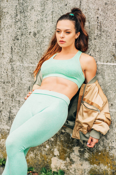 Outdoor portrait of young beautiful fit woman, wearing green activewear, athlete model posing next to grey urban background, leaning on the wall, sport fashion  - Photo, Image