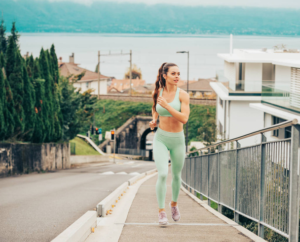 Portrait of young beautiful fit woman running up the hill, wearing green activewear, athlete model posing outdoors, sport fashion - Photo, Image