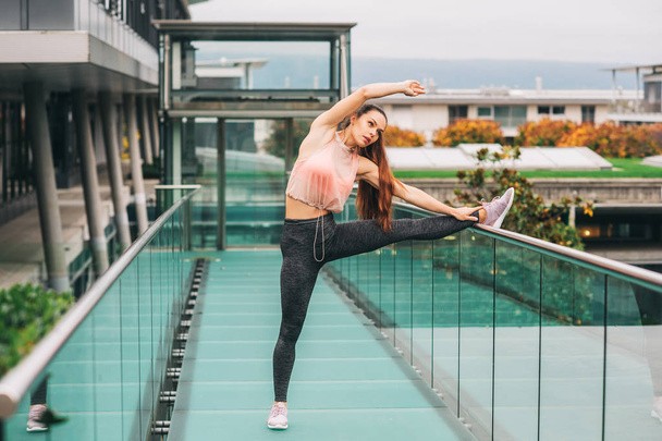 Outdoor portrait of young beautiful fit woman stretching legs, wearing fashionable activewear, athlete model posing in urban background, sport fashion - 写真・画像