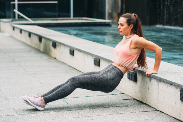 Outdoor portrait of young beautiful fit woman doing tricep dips, wearing fashionable activewear, athlete model posing in urban background, sport fashion - Photo, Image
