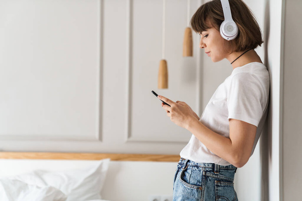 Amazing young lady indoors at home listening music with headphones using mobile phone. - Foto, afbeelding