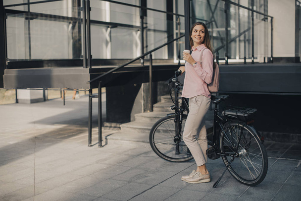 Güzel genç bayan bisikletçi şehir ortamında elektrikli bisikletin yanında bir fincandan kahve içer. - Fotoğraf, Görsel