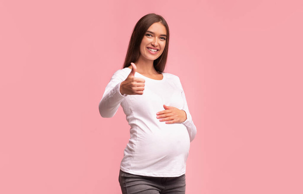 Positiivinen raskaana oleva nainen elehtii peukalot pystyssä yli vaaleanpunainen tausta
 - Valokuva, kuva