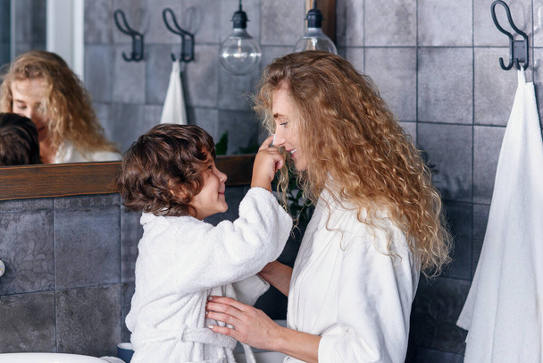 Little handsome son puts skin care cream on his beautiful mothers face. Pretty mom and her cute son dressed in white bathrobes stand together in modern stylish bathroom. - Foto, immagini