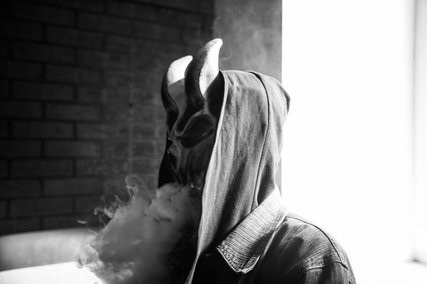 Fatal vaping activity. Vape man in devil mask and a hood smoking an electronic cigarette indoors. Puffs of steam. Bad habit that is harmful to health. Vaping activity. Black and white. - Фото, зображення