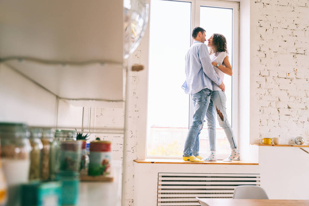 Jovem casal feliz abraçando no peitoril da janela em seu apartamento
, - Foto, Imagem