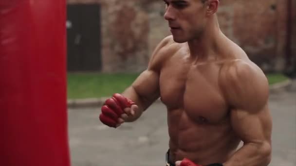 Bezkoszulkowy bokser bokserski uderzający w worek treningowy podczas treningu na świeżym powietrzu. Zbliżenie - Materiał filmowy, wideo