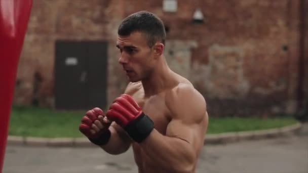 Slow motion. Close view. Male shirtless boxer hitting a big punching bag with his fists during street workout - Footage, Video