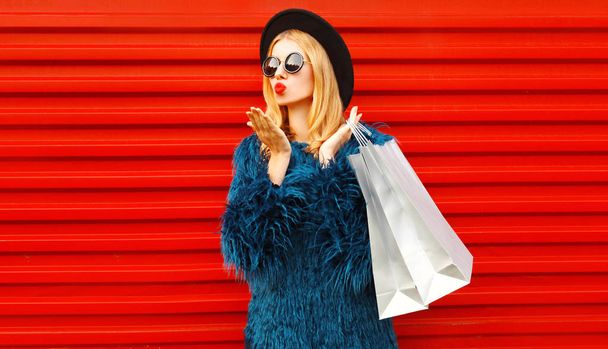 Side view beautiful woman with shopping bags blowing red lips sending sweet air kiss, stylish female model wearing blue faux fur coat, round hat and sunglasses over red wall background - Foto, Imagem
