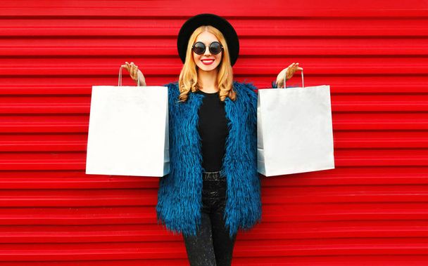 Stylish woman showing two same shopping bags wearing blue faux fur coat, round hat and sunglasses over red wall background - Fotó, kép
