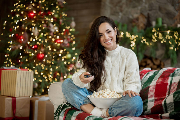 happy brunette woman wearing sweater watching tv sitting on the sofa with popcorn in room decorated for celebrating new year and christmas eve - Zdjęcie, obraz