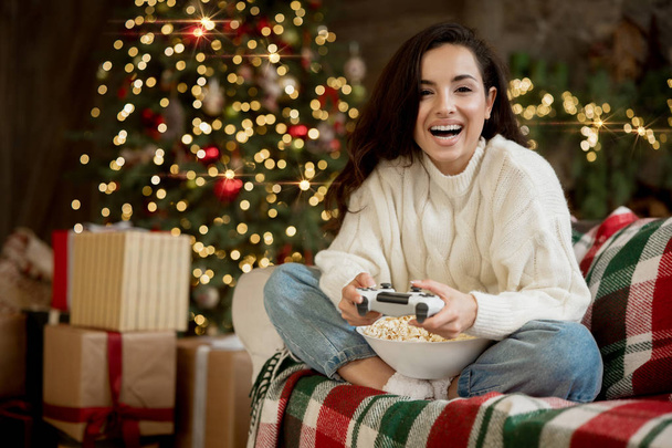 brunette woman wearing sweater playing video game on the sofa with popcorn in room decorated for celebrating new year and christmas festive mood - Foto, imagen