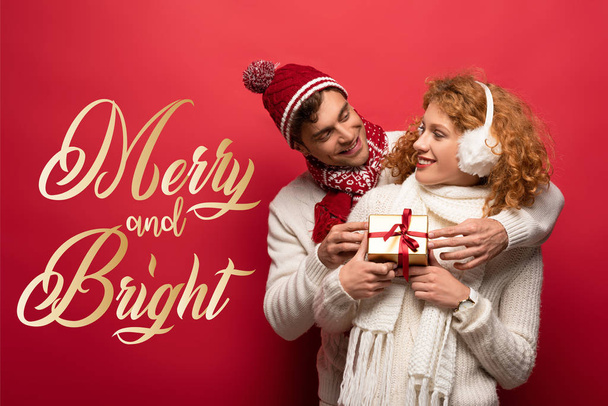happy couple in winter outfit holding christmas gift on red with merry and bright lettering - Foto, Bild