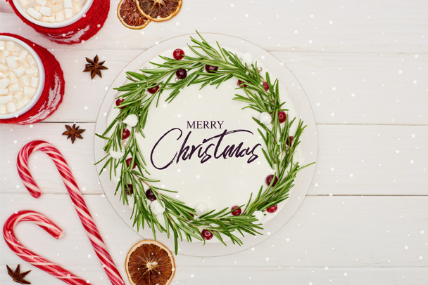 vista dall'alto di bastoncini di zucchero, due tazze di cacao e torta di Natale con allegra illustrazione natalizia su tavolo di legno bianco
  - Foto, immagini