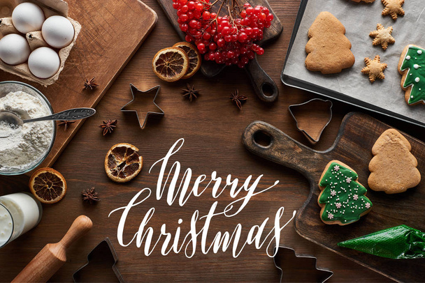 bovenaanzicht van geglazuurde kerstkoekjes bij ingrediënten, deegvormen en viburnum op houten tafel met vrolijke kerstillustratie - Foto, afbeelding