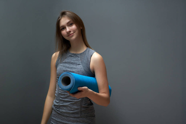 Athletic woman with blue rug in hands on background of gray wall - Foto, Imagen