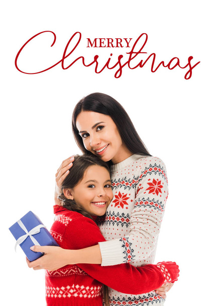 cheerful mother hugging daughter and holding preset isolated on white with merry Christmas lettering - Фото, изображение