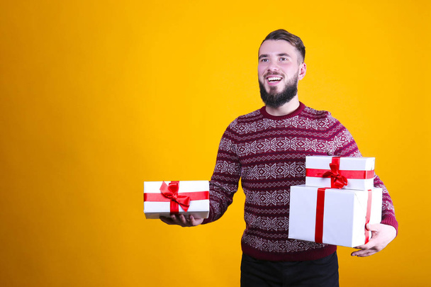 Studio portrait of handsome bearded man wearing christmas sweater with snowflake ornament holding presents, posing over the yellow wall, copy space. Festive background. Male with facial hair smiling. - Photo, Image