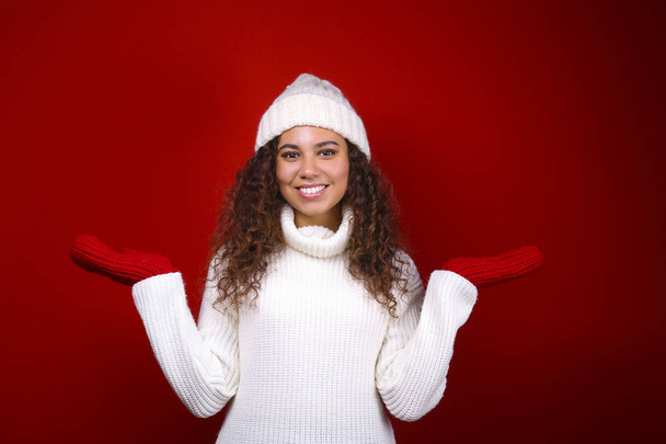 Studio portrait of young woman with dark skin and long curly hair wearing knitted turtle neck sweater over the festive red wall with a lot of copy space for text. Close up, isolated background. - Foto, immagini