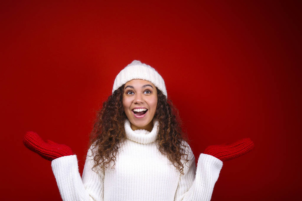 Studio portrait of young woman with dark skin and long curly hair wearing knitted turtle neck sweater over the festive red wall with a lot of copy space for text. Close up, isolated background. - Fotó, kép