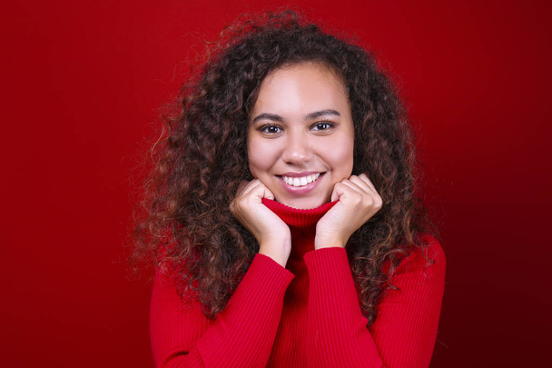 Studio portrait of young woman with dark skin and long curly hair wearing knitted turtle neck sweater over the festive red wall with a lot of copy space for text. Close up, isolated background. - Foto, Bild
