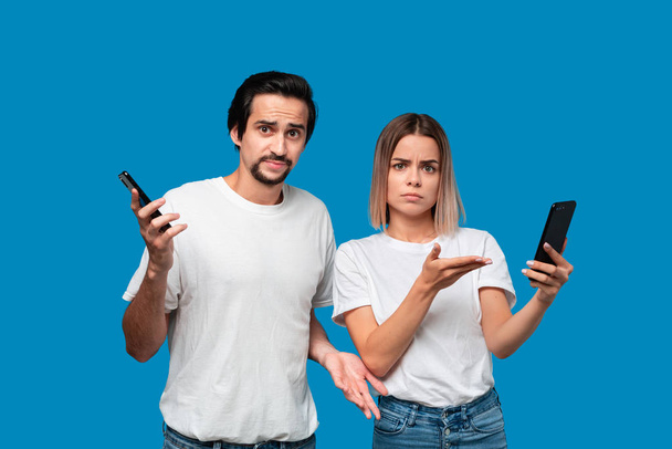 Brunet man in witte tees en blauwe jeans is geschokt te kijken naar het smartphone scherm van een blonde vrouw - Foto, afbeelding