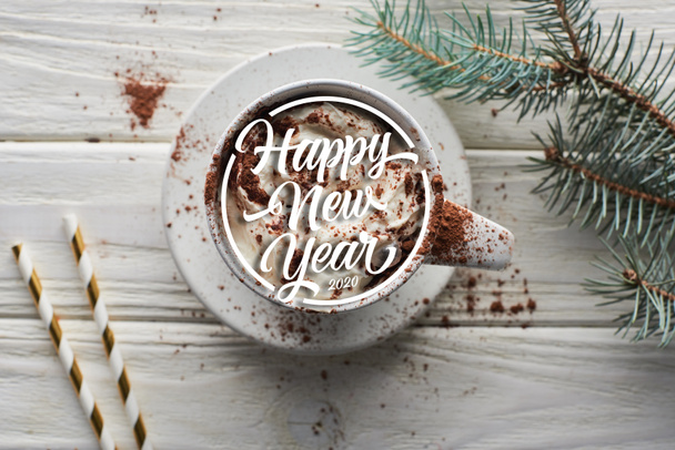 vrchní pohled na vánoční kakao s kakaovým práškem a šlehačkou v hrnku na bílém dřevěném stole u jedlové větve se šťastným novoročním nápisem - Fotografie, Obrázek