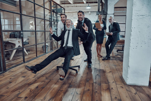 Playful businessmen pushing their boss on the office chair while running in the hallway              - Zdjęcie, obraz