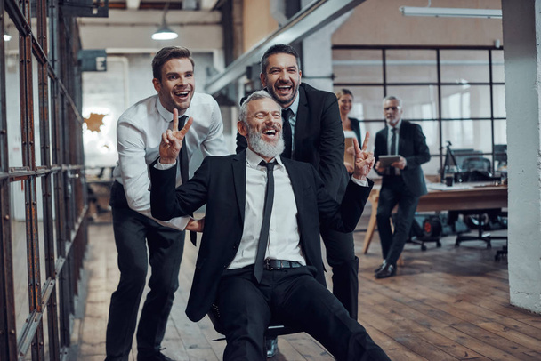 Cheerful businessmen pushing their boss on the office chair while running in the hallway - Zdjęcie, obraz