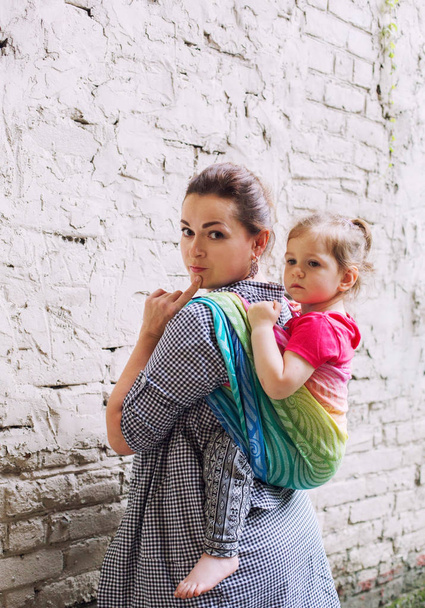 Matka i maluch córka wygląd rodziny za pomocą owinąć nosidełko proca szalik z powrotem chodzić w mieście, pobyt w pobliżu białej ściany, mobilność i nowoczesne koncepcji rodzicielstwa - Zdjęcie, obraz