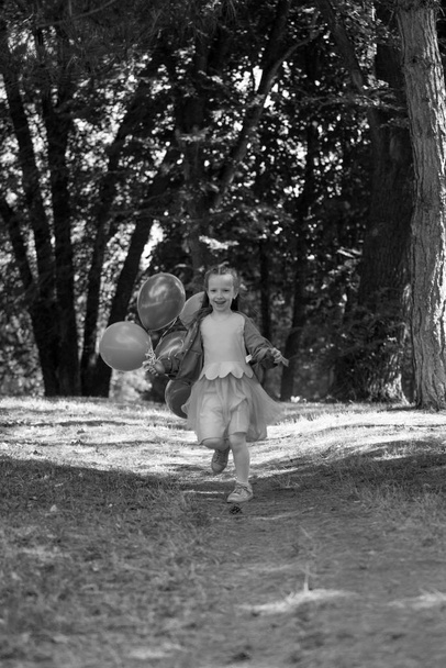 Μικρό χαριτωμένο κορίτσι τρέχει στο πάρκο με ένα μάτσο μπαλόνια. Γελάει προκλητικά. Η έννοια της ευτυχισμένης παιδικής ηλικίας. - Φωτογραφία, εικόνα