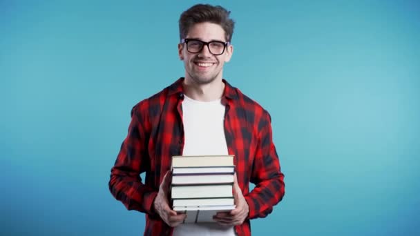 European student in red plaid shirt on blue background in studio holds stack of university books from library. Copy space. Guy smiles, he is happy to graduate. - Záběry, video