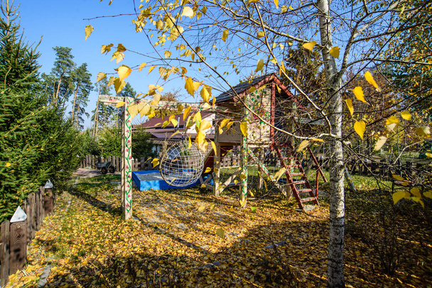 La tierra del patio está cubierta de hojas doradas. Jardín delantero de una casa con árboles de follaje dorado. El abedul en primer plano está cubierto con follaje dorado. Zona de juegos
. - Foto, imagen