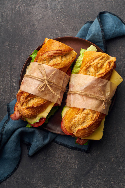 Fresh big baguette sandwiches with bacon and chedder cheese and lettuce with vegetables on textile  - Photo, image