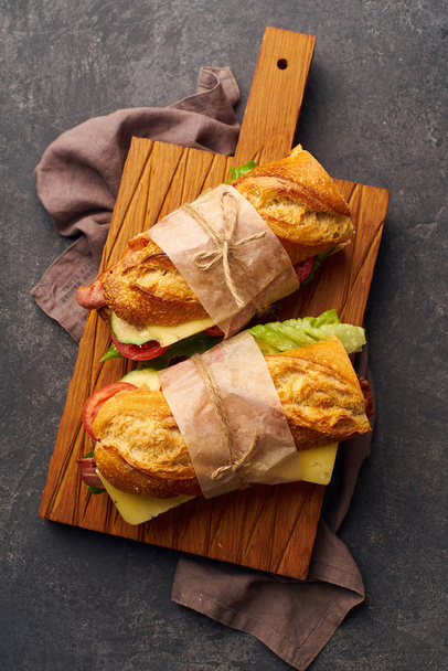 Fresh big baguette sandwiches with bacon and chedder cheese and lettuce with vegetables on wooden cutting board  - Φωτογραφία, εικόνα