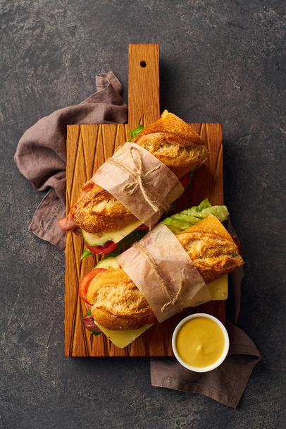 Fresh big baguette sandwiches with bacon and chedder cheese and lettuce with vegetables on wooden cutting board  - Фото, изображение