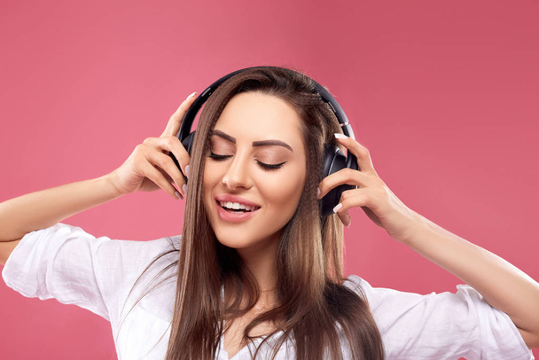 Красивая молодая женщина в беспроводных наушниках слушает музыку с помощью мобильного телефона и танцует на розовом фоне. Девушка использует беспроводные наушники
 - Фото, изображение