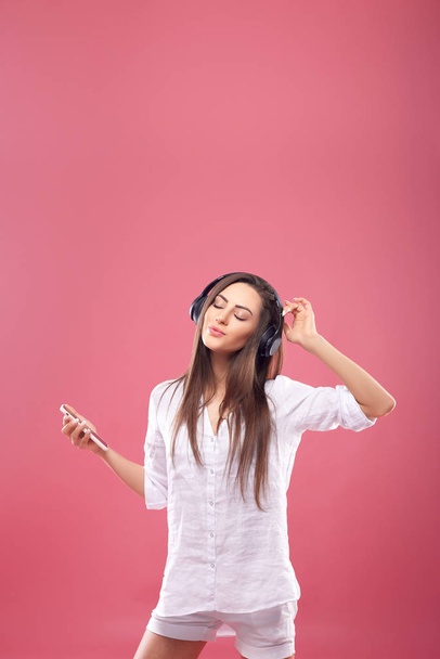 Beautiful young woman in wireless headphones listening to music using mobile phone and dancing on pink background. Girl uses wireless earphones - Fotografie, Obrázek