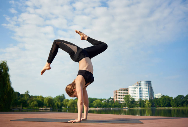Mujer practica yoga en parque de la ciudad haciendo handstand cerca de lago contra fondo de edificios de gran altura bajo cielo azul
  - Foto, imagen