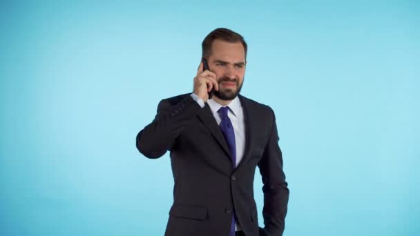 Unsatisfied businessman speaks on phone and expresses his disagreement isolated on blue studio background. Denying, rejecting, disagree, portrait of handsome man. 4k. - Video, Çekim