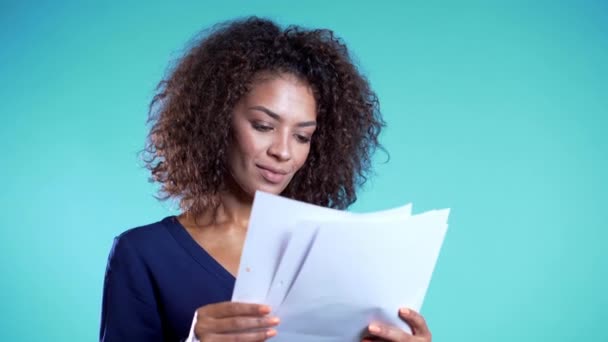 Pretty african american female boss checks documents, utility bills. Serious businesswoman standing on blue background. She is satisfied with work of staff. 4k - Séquence, vidéo