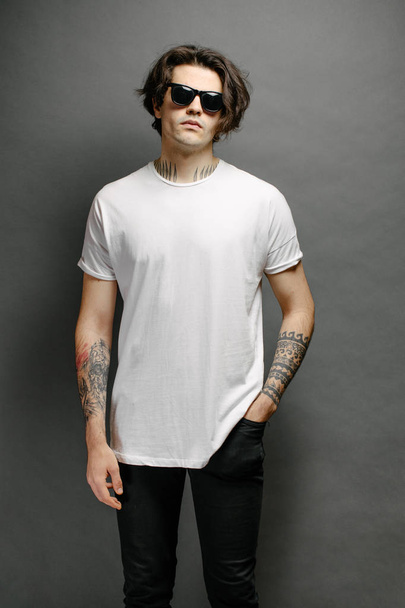 Hipster handsome male model with glasses wearing white blank t-shirt and black jeans with space for your logo or design in casual urban style - Foto, Bild