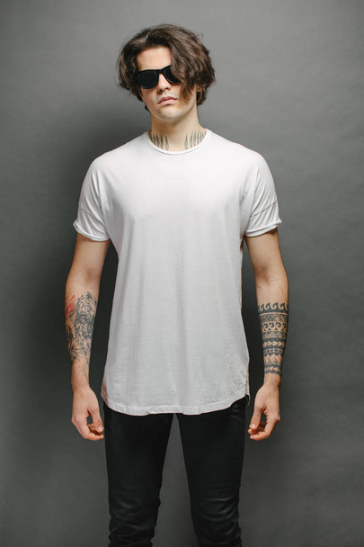 Hipster handsome male model with glasses wearing white blank t-shirt and black jeans with space for your logo or design in casual urban style - Foto, Bild