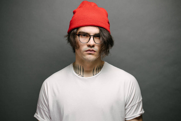 Hipster handsome male model with glasses wearing white blank t-shirt and red pants and a red hat with space for your logo or design over gray background - Fotó, kép